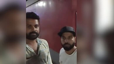 Two men with beards and dark hair standing indoors against a red wall.