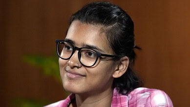 Young woman with glasses smiling, wearing a pink checkered shirt, indoor setting.