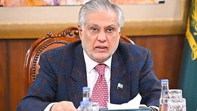 Pakistani official speaking at a press conference with flags in the background.
