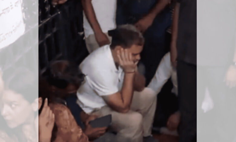 Man sitting on the ground with hand on face, surrounded by people at a protest or gathering.