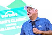 Workday CEO speaking at charity event with a microphone, wearing a blue polo shirt and cap.
