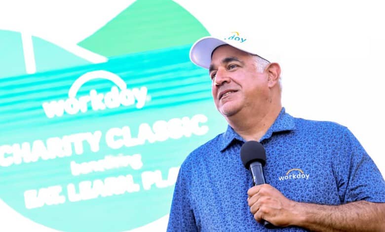 Workday CEO speaking at charity event with a microphone, wearing a blue polo shirt and cap.