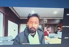 Man with beard and dark jacket in office setting with two colleagues in background.