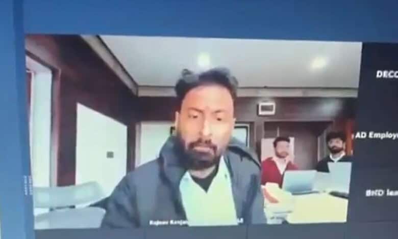 Man with beard and dark jacket in office setting with two colleagues in background.