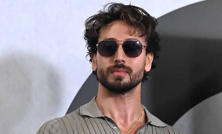 Man with curly hair, beard, sunglasses, wearing a grey shirt, against a plain background.