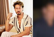 Portrait of a man with curly hair and a beard, wearing a patterned shirt, sitting indoors.