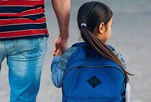 Young girl with a blue backpack holding hands with an adult, walking outdoors.