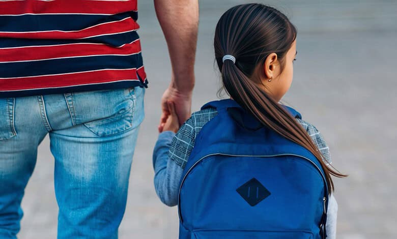 Young girl with a blue backpack holding hands with an adult, walking outdoors.