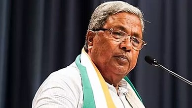 Former CM Siddaramaiah speaking at a press conference with a microphone and a dark background.