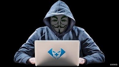Person in hoodie wearing Guy Fawkes mask using a laptop with hacker logo.