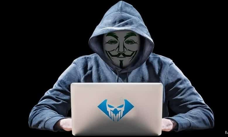 Person in hoodie wearing Guy Fawkes mask using a laptop with hacker logo.