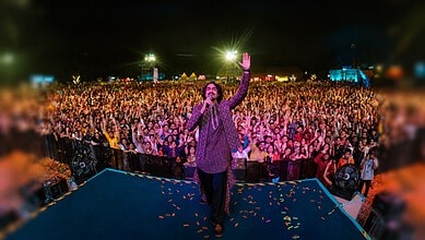 A speaker addresses a large crowd at night during a public event with bright lights and tents in the背景.