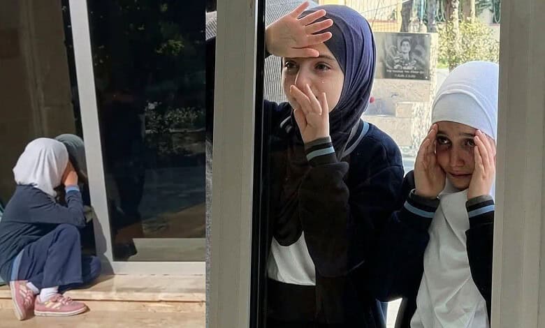 Girls wearing headscarves look out from school window, appearing worried and anxious.