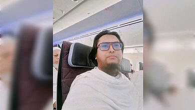 Man with glasses sitting on an airplane, wearing a white shawl, looking forward.
