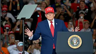 Former US President Donald Trump speaking at a rally, wearing a red 'USA' cap and blue suit.