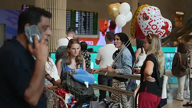 People waiting at airport with balloons, luggage, and a woman with a backpack.