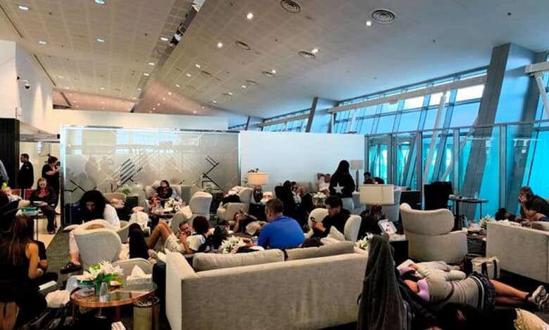 Passengers waiting at airport lounge with seating and large windows in the background.