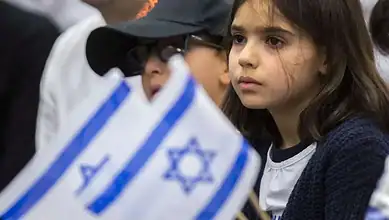 Young girl with serious expression holding an Israeli flag at a public event.