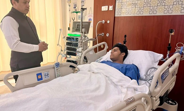 Young male patient lying in hospital bed with medical equipment, visitor standing nearby.