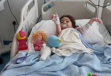 Young girl lying in hospital bed with toys, connected to medical equipment, recovering from treatment.