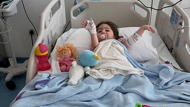 Young girl lying in hospital bed with toys, connected to medical equipment, recovering from treatment.
