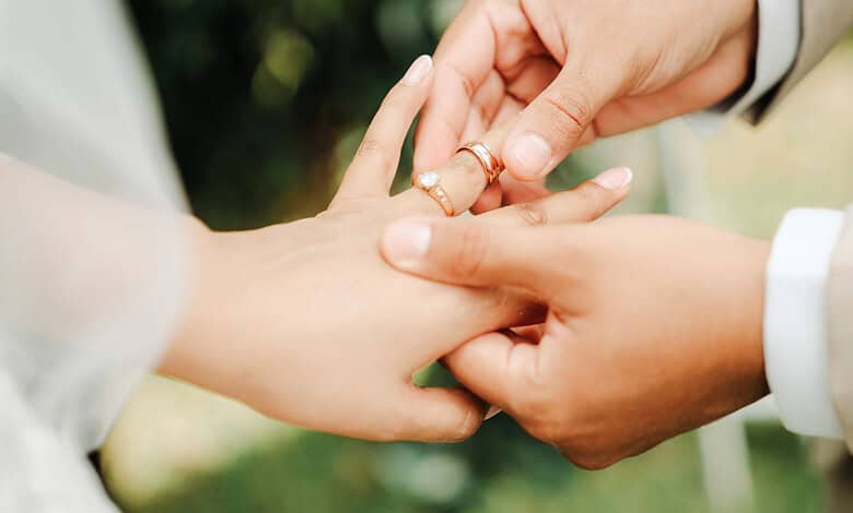 Close-up of a couple holding hands, wedding rings visible, outdoors setting.