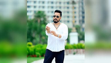 Mika Singh wearing sunglasses and a white shirt, standing outdoors with a cityscape background.