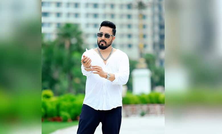Mika Singh wearing sunglasses and a white shirt, standing outdoors with a cityscape background.