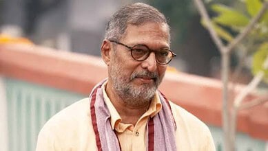 Nana Patekar smiling outdoors with glasses and a beard, wearing a beige shirt and scarf.