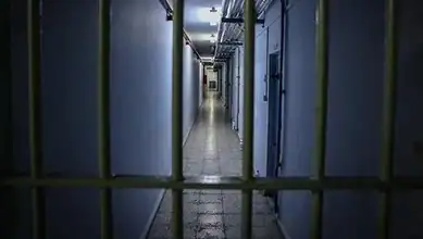 Empty prison corridor with barred cells and dim lighting.