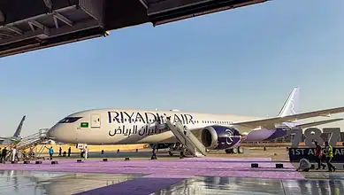 Riyadh Air aircraft parked at the airport with stairs and ground crew nearby.