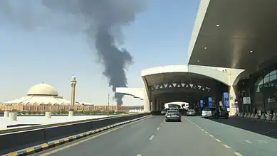 Smoke rising from Riyadh airport after attack, with vehicles on the road in the foreground.