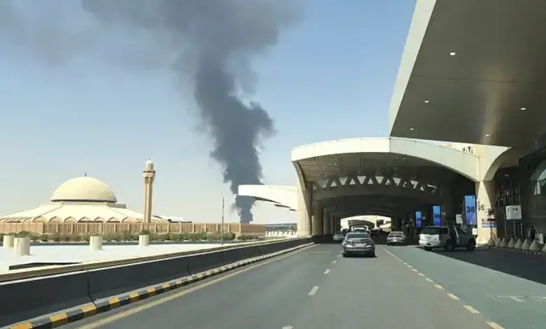 Smoke rising from Riyadh airport after attack, with vehicles on the road in the foreground.