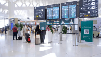 Passengers inside an airport terminal near departure information screens and check-in counters.