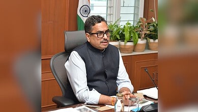 Indian official in formal attire seated at desk with plants and Indian flag in background.