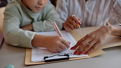 Children and adult working on homework with pens and papers at a table.
