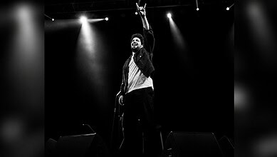 Samay Raina performing on stage with microphone, raising hand, in black and white.