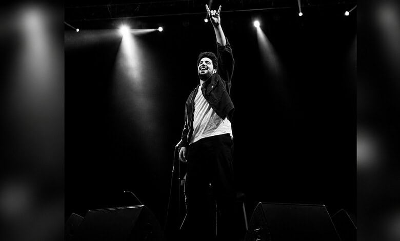 Samay Raina performing on stage with microphone, raising hand, in black and white.
