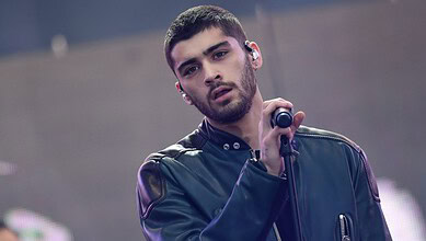 Zayn Malik singing on stage with a microphone, wearing a dark jacket and looking into the distance.